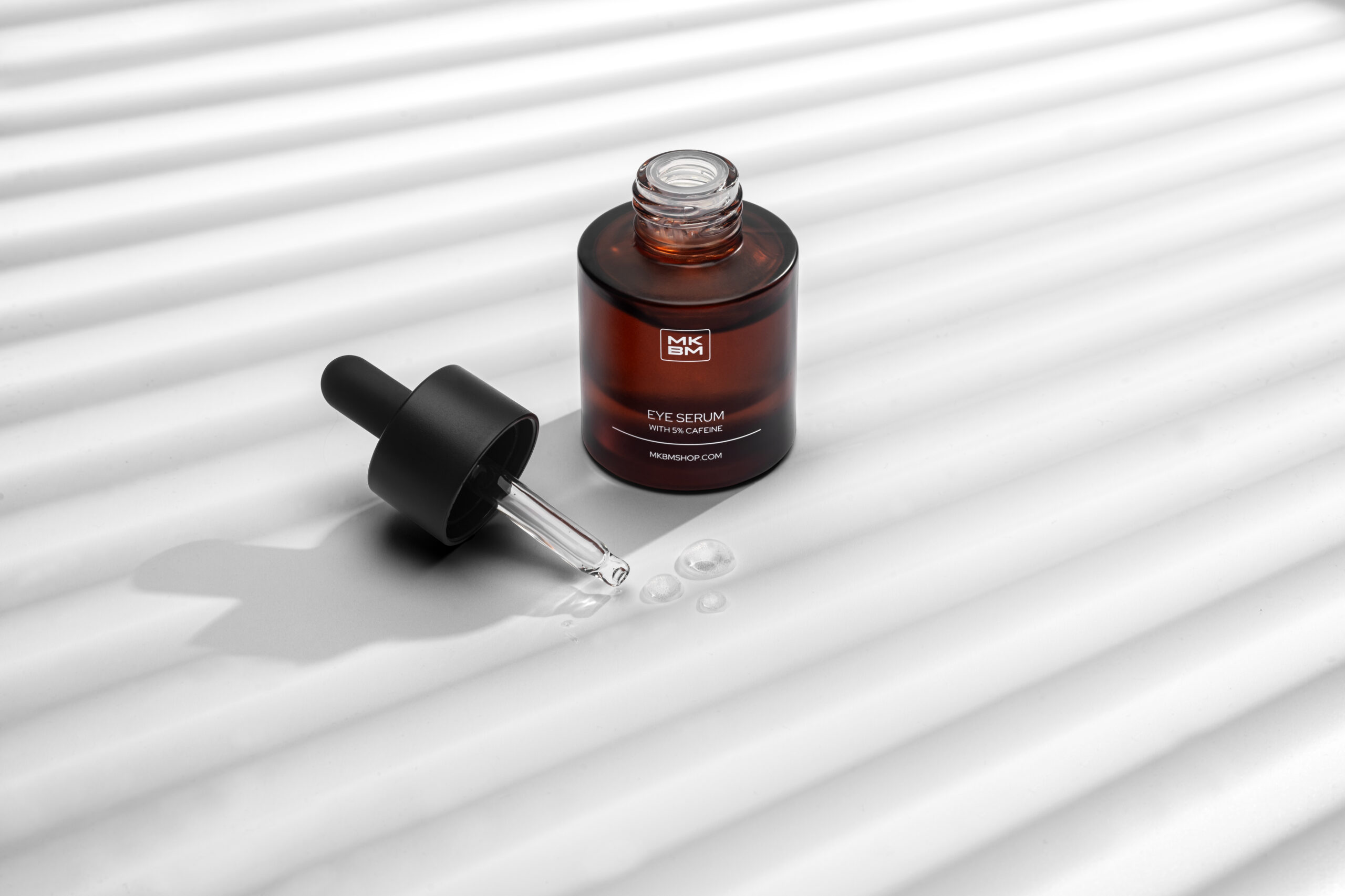 Creatieve skincare productfotografie van MKBM Eye Serum met pipet en serumdruppels op witte achtergrond met grafische schaduwen.