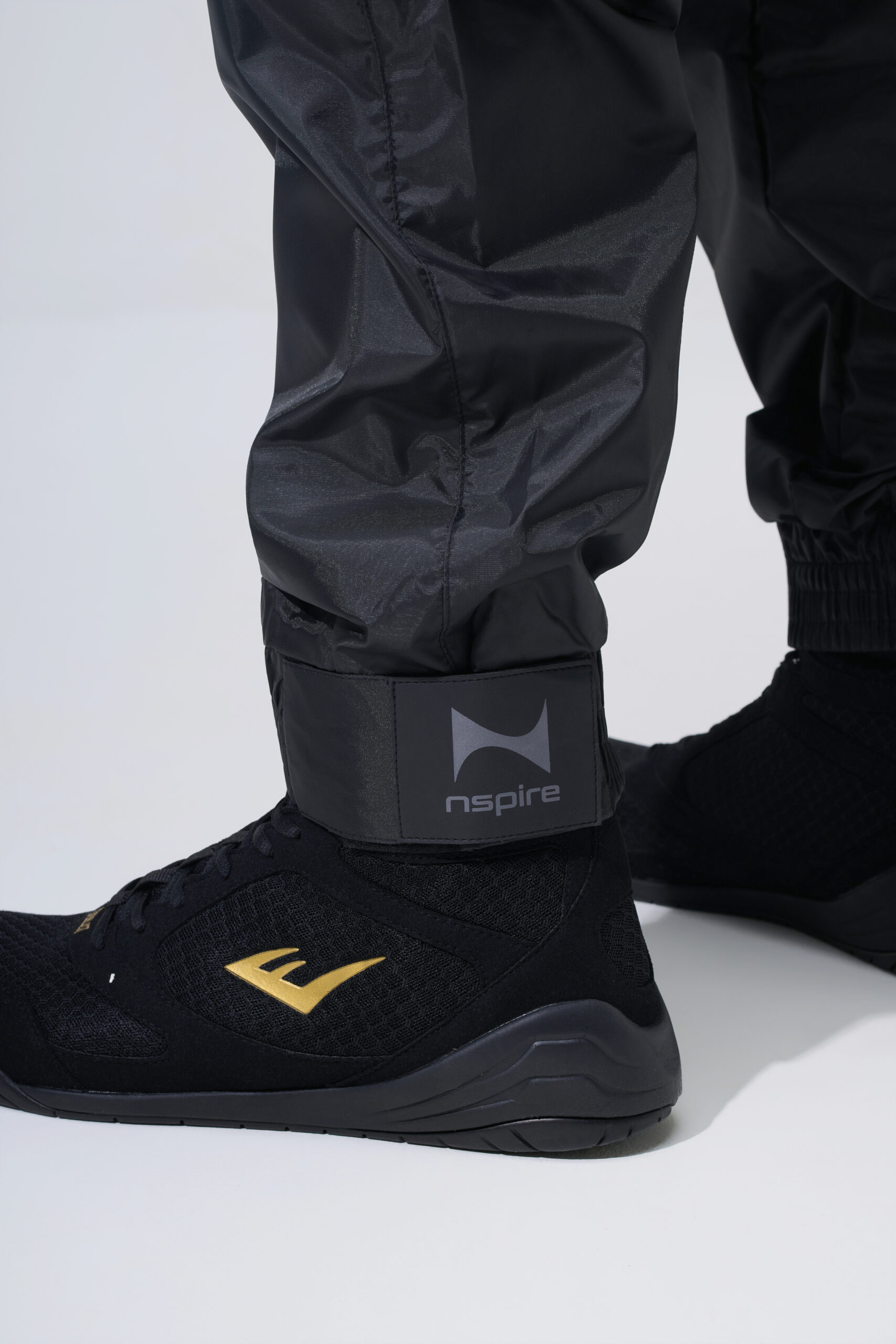 Close-up productfotografie van zwarte Nspire trainingsbroek en sportschoen met merkdetail rond de enkel.