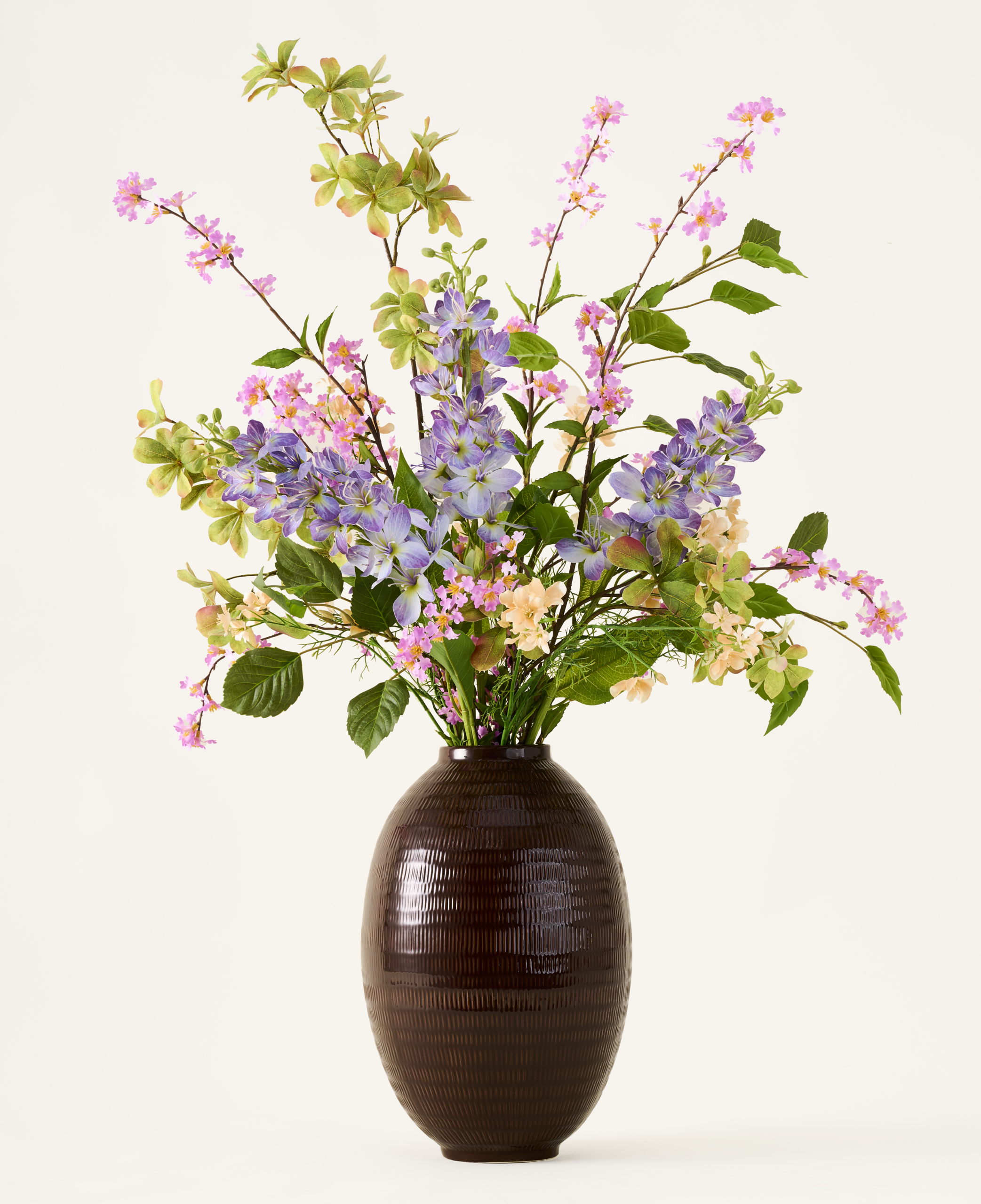 Productfoto van een pastel kunstbloemenarrangement met paarse hortensia en roze bloesemtakken in een donkerbruine glazen vaas.