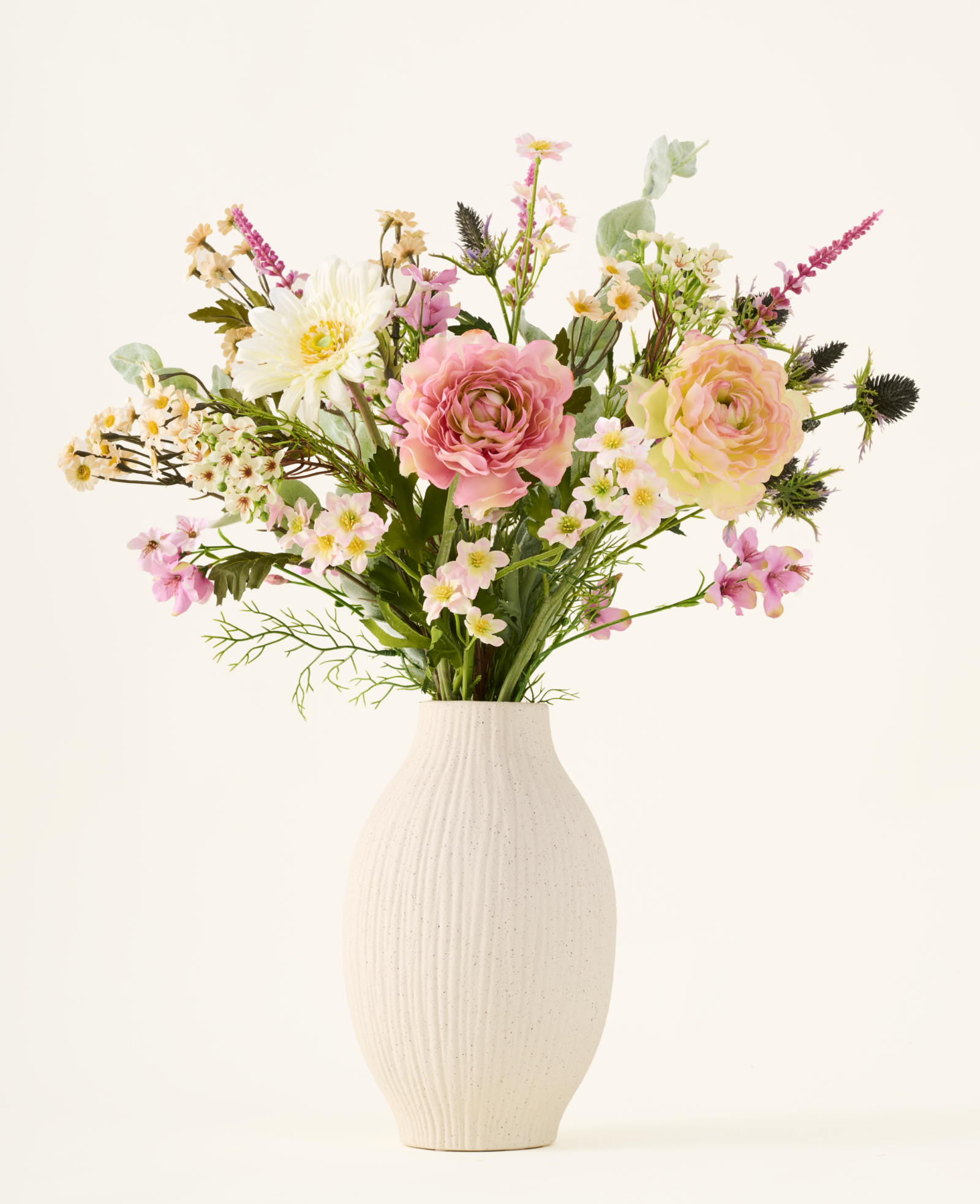 Lichte studiofoto van een romantisch kunstbloemenboeket met roze ranonkels, witte bloemen en groen in een crèmekleurige vaas.