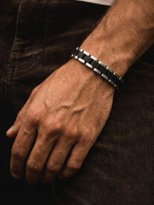 Close-up van een zwart-zilveren herenarmband met geometrisch patroon, gedragen om de pols en sfeervol gefotografeerd tegen donkere kleding.