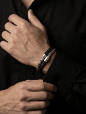Dubbele, donker gevlochten herenarmband met zilverkleurige sluiting, gedragen bij een zwarte blouse in een stijlvolle, donkere setting.